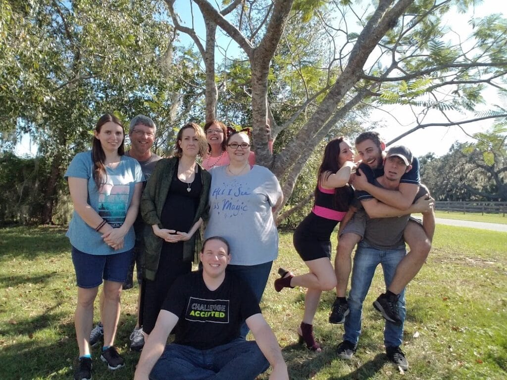this is a picture of my whole family outside under the tree posing for the picture and being goofy too. our friend has my son on his back and my daughter is leaning up against him. My other daughter is very pregnant and we're all smiling. there is a bunch of us.