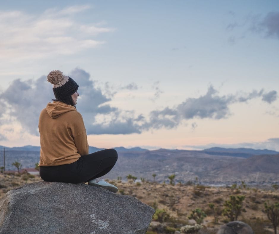 You are stronger than you think. woman sitting on rock overlooking valley. she has on a tan sweater and a hat, you can only see the back of her with the clouds in the distance.