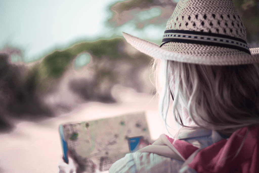 blonde woman in a hat facing away from the camera looking at a map. there's a road in front of her.