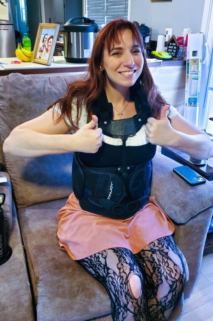 Wrecked Car. Daughter in Hospital Bed. Nicole in her body brace after the car accident. She's got brown hair and has on a white shirt with a pink skirt and lace leggings underneath. Shes smiling and has 2 thumbs up for the camera. We're so grateful she's alive.