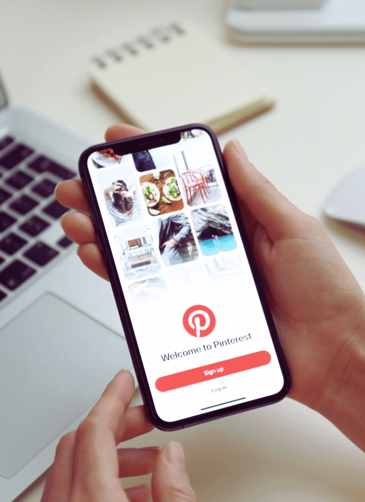 how to make money on pinterest. theres a womans hands holding a smartphone with a pinterest screen on the cover. there are words that say welcome to pinterest. and there is a keyboard in the background.