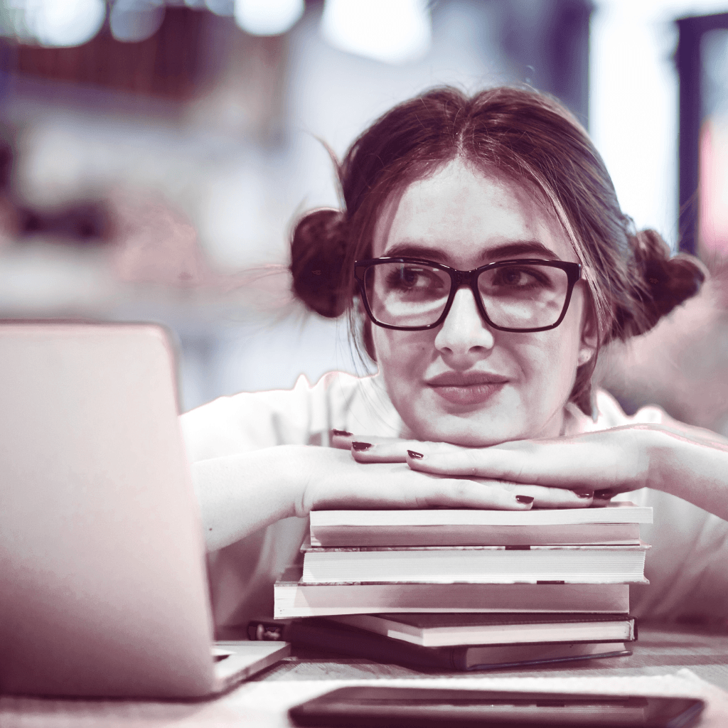 girl with glasses dreaming on stack of books in front of laptop. she's smiling and has on glasses with brown hair and her hair is up in two buns on either side of her head.