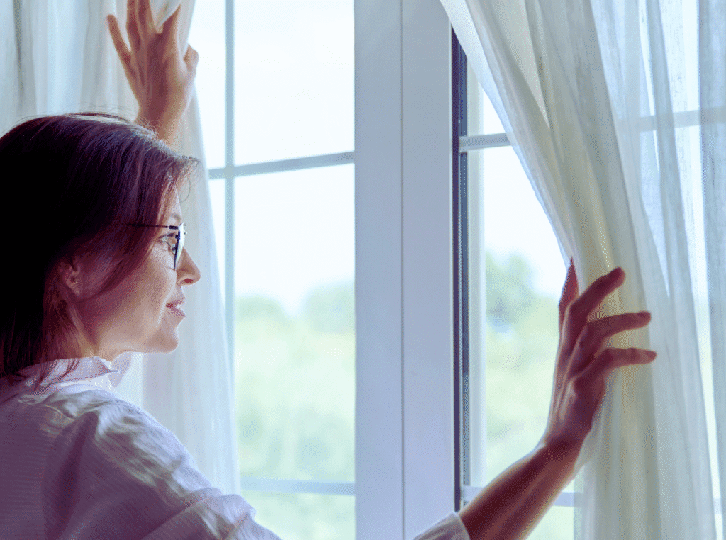 Bible verses about forgiveness. older woman with glasses pulling curtains open and looking out window during the day.