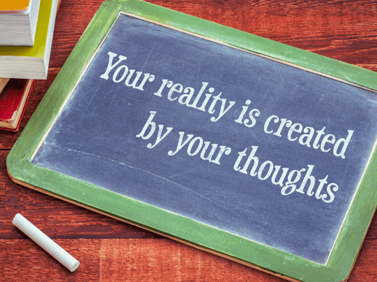 Be Careful Of Your Thoughts Because, image of a small hand held chalkboard with a piece of chalk under it. The writing on the board says: your reality is created by your thoughts.