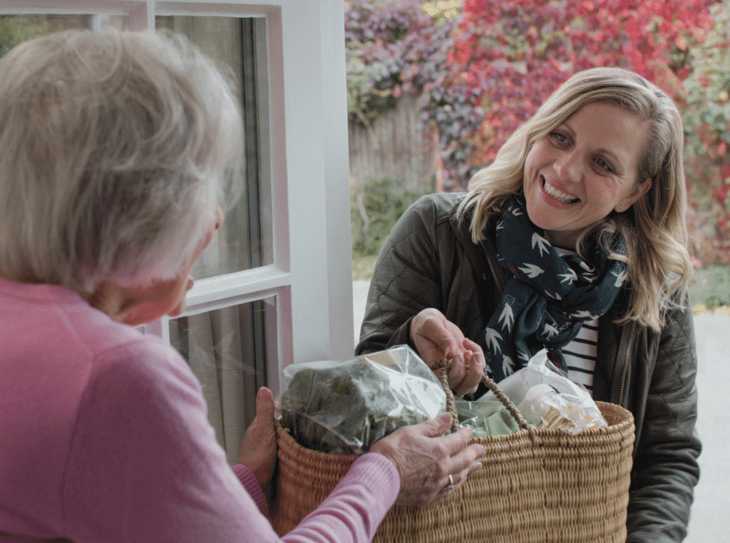 Good Values And Morals. tired woman at the door handing a basket of food to an elderly woman that needs help. both are smiling.