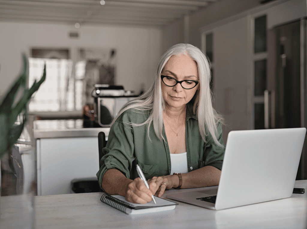 Example Of Keyword Stuffing. older woman at laptop with a notebook and pen next to her and she's writing something like taking notes.