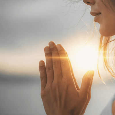 prayers for adult children. woman with her eyes closed, hands in prayer, praying outside in front of the sunset.