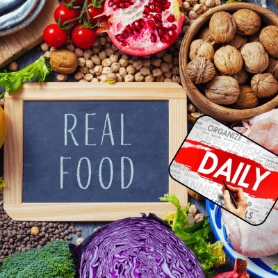 real food daily. a mixture of different real foods laying on a table. nuts, pomegranites, red cabbage, broccoli, etc. and a little chalkboard in the center that says: real food. and off to the side it says: daily.