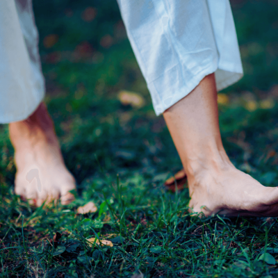 What is earthing and grounding? Woman walking barefoot on the ground so her feet are touching the grass. She's wearing blue pants.